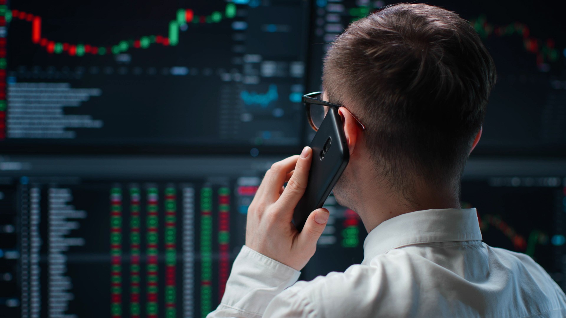Back view of trader working with multiple computer screens full of charts and data talking on phone