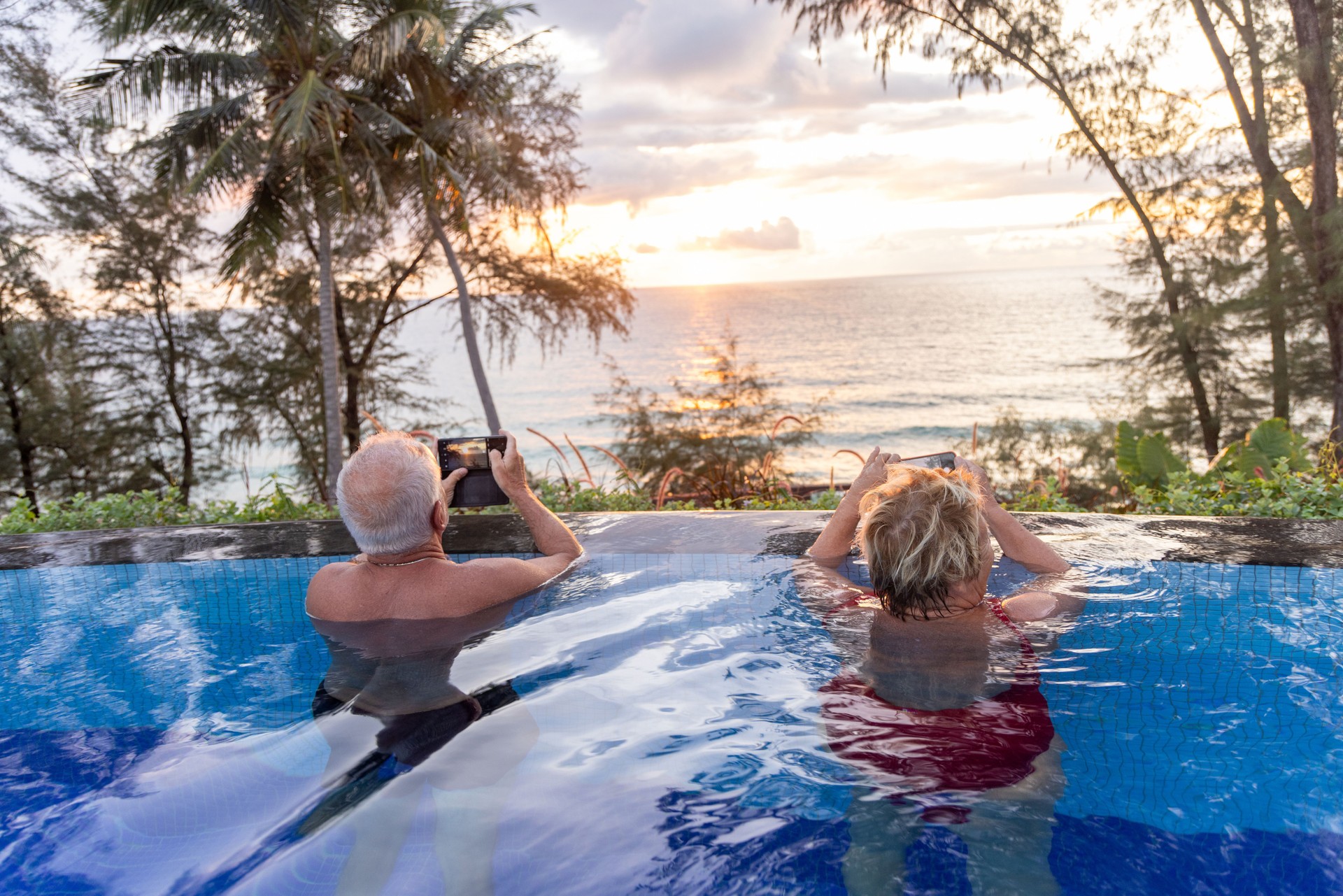 Two seniors doing social media from swimming pool, they photograph the sunset